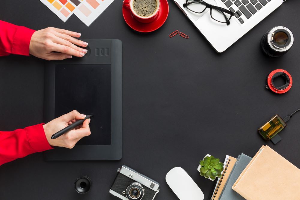 Creative workspace of a digital artist using a drawing tablet, surrounded by tools including a laptop, red coffee mug, color swatches, camera gear, and design accessories on a clean desk. Perfect for content related t